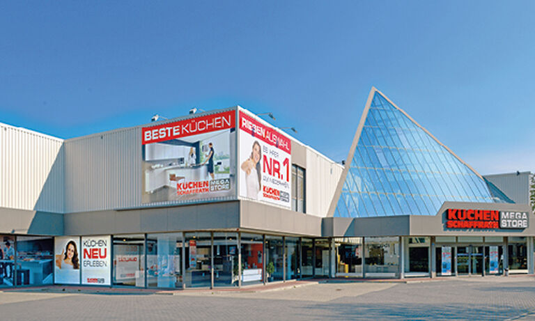 Das Bild zeigt den „Küchen Schaffrath Mega Store“ mit einer modernen Architektur, die durch eine auffällige, gläserne Pyramidenstruktur auf der rechten Seite hervorsticht. Auf der linken Gebäudeseite sind große Werbebanner angebracht, die die „BESTE KÜCHEN“ und eine „RIESEN AUSWAHL“ an Küchen bewerben. Einer der Schriftzüge hebt hervor: „NR. 1 in Küchenkompetenz“. Darunter befinden sich Glasfenster, die einen Blick in den Ausstellungsraum ermöglichen. Der Eingang ist zentral unter einem flachen Vordach mit dem Schriftzug „KÜCHEN SCHAFFRATH MEGA STORE“ in roten und weißen Buchstaben. Der Vorplatz ist gepflastert, und der Himmel ist strahlend blau, was das Gebäude einladend und freundlich wirken lässt.